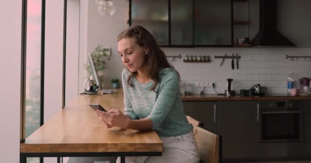 Mujer joven sentarse en la cocina doméstica utilizando el teléfono inteligente — Vídeos de Stock