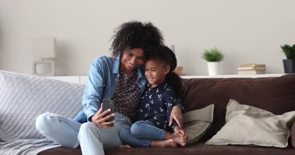 Mãe africana e pequena filha sentam-se no sofá usar smartphone — Vídeo de Stock