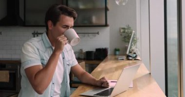 Adam mutfakta oturur, kahve içer ve dizüstü bilgisayarla çalışır.