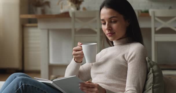 Mujer pacífica relajándose en el sofá sosteniendo libro de lectura de la taza — Vídeo de stock