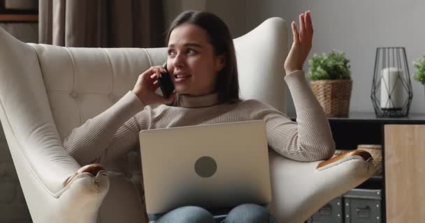 Mujer sentada en sillón con portátil hablando por teléfono — Vídeos de Stock