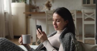 Ekoseli çay fincanı ile kaplı evde dinlenen kadın akıllı telefon kullanıyor.