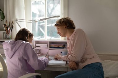 Odaklanmış genç bir anne, genç bir kıza ödevlerinde yardım ediyor..