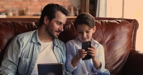 Papá e hijo pequeño hablando sosteniendo gadgets sentarse en el sofá — Vídeos de Stock