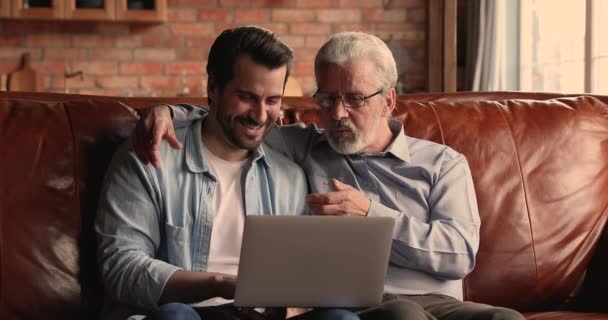 Uomo più anziano suo figlio millenario sedersi sul divano con il computer portatile — Video Stock