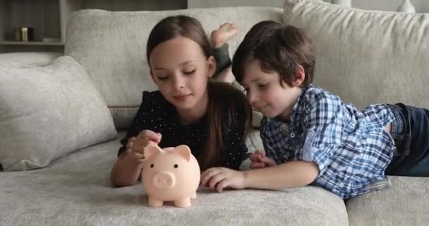 Kleine kinderen die op de bank liggen en munten in de spaarpot gooien — Stockvideo