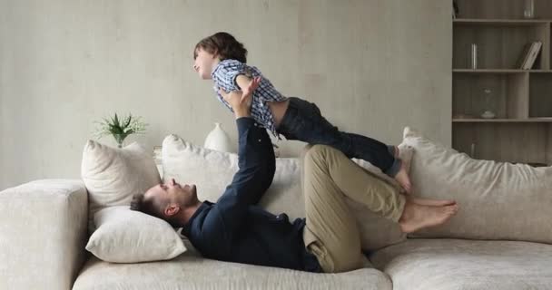 Отец лежит на диване лифты на протянутых руках маленький сын — стоковое видео