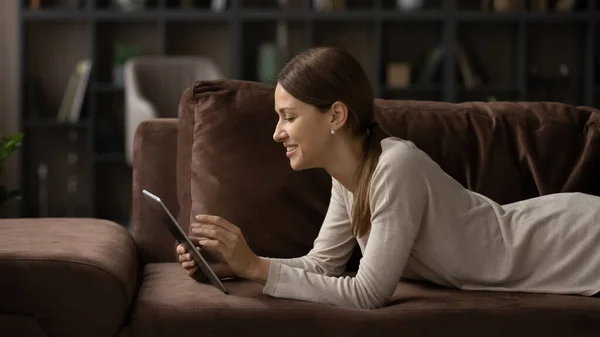 Mladá dáma odpočívat na gauči svitek novinky na tablet on-line — Stock fotografie