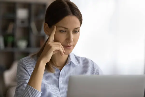 Вдумчивая молодая женщина читает документ на экране компьютера на рабочем месте — стоковое фото