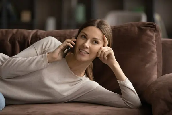 Усміхнена тисячолітня жінка лежить на зручному дивані, розмовляючи по телефону — стокове фото