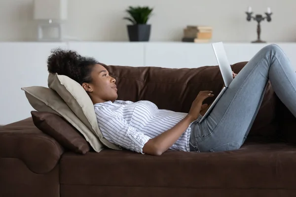 Relaxado despreocupado jovem mulher africana usando computador, deitado no sofá. — Fotografia de Stock