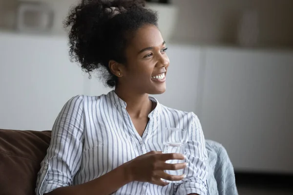 Smiling dreamy young african woman holding glass of mineral water. — Stock Photo, Image