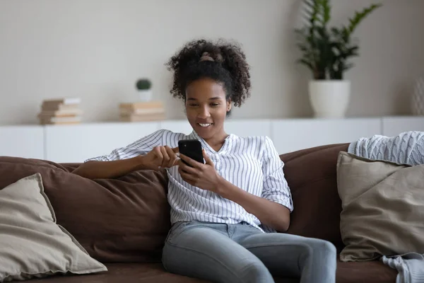 Ontspannen jonge Afrikaanse vrouw met behulp van smartphone toepassingen thuis. — Stockfoto