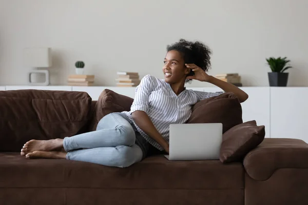 Dromerige duizendjarige Afrikaanse vrouw rusten op de bank met computer. — Stockfoto