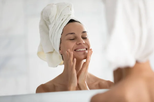 Happy woman with towel on head touching face at mirror — Stock Photo, Image