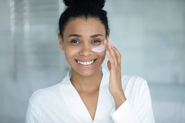 Mujer feliz en albornoz aplicando máscara para el tratamiento de la piel debajo de los ojos —  Fotos de Stock