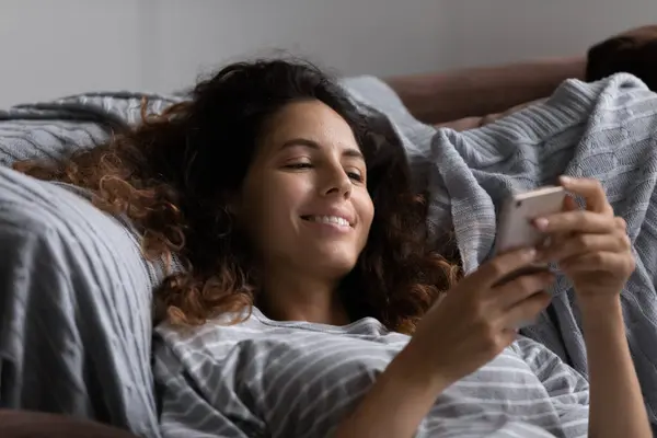 Sorridente donna ispanica rilassarsi a letto con il cellulare — Foto Stock