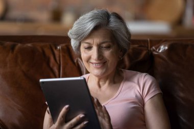 Olgun, gülümseyen bir kadın evinde tabletle oturuyor.
