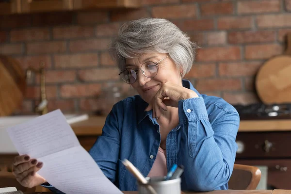 Ältere Frau sitzt in Küche am Tisch und liest Papier — Stockfoto