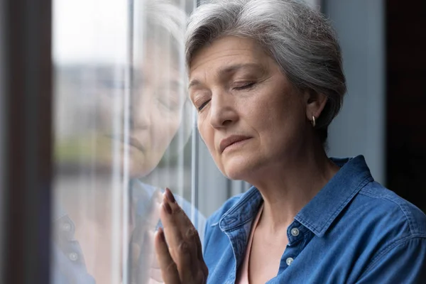 Triste donna anziana depressa in piedi vicino alla finestra con gli occhi chiusi — Foto Stock