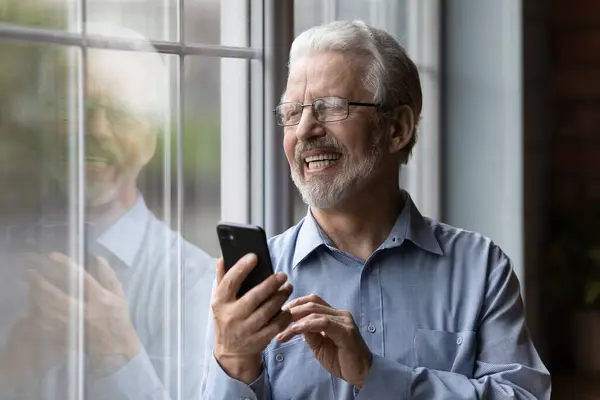 Hombre mayor alegre sosteniendo teléfono inteligente mirando por la ventana —  Fotos de Stock