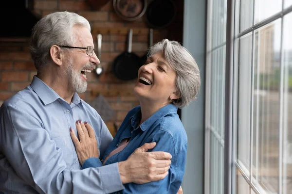 Äldre gråhåriga morfar kram mormor skrattar stående inomhus — Stockfoto