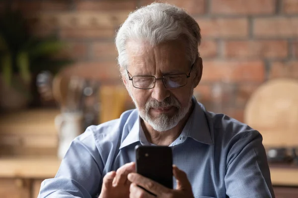 Hombre maduro serio en gafas sentarse en la cocina sosteniendo teléfono inteligente — Foto de Stock