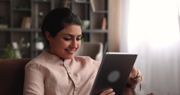 조심스럽게 웃고 있는 인도 여자가 태블릿 장치를 가지고 소파에 누워 있다 — 비디오