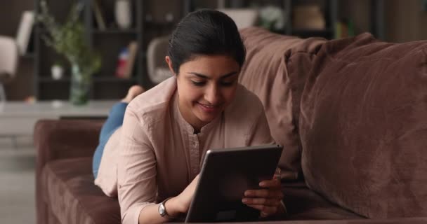 Hintli kadın kanepeye uzanıp dijital tablet kullanarak eğleniyor. — Stok video