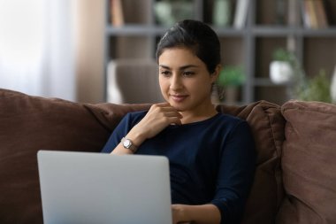 İlgilenen Hintli genç bayan dizüstü bilgisayar kullanarak kanepeye uzanıyor.