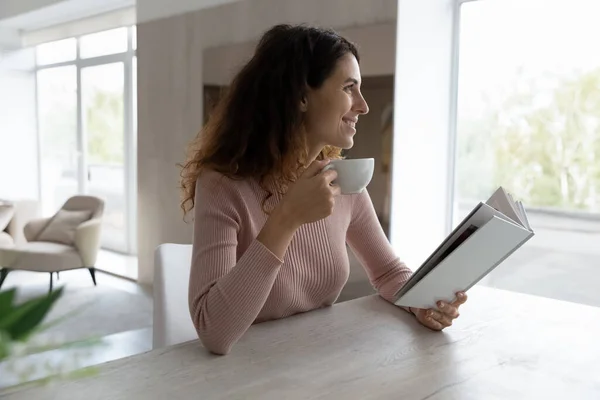 Gülümseyen Latin kadın kahve içiyor evde kitap okuyor. — Stok fotoğraf
