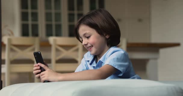 Хлопчик сидить на дивані зі смартфоном, розважаючись грати в ігри — стокове відео