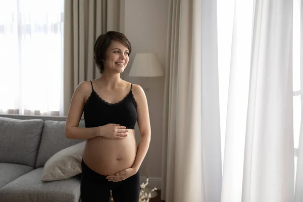 Usmívající se zasněná těhotná žena dotýkající se břicha, dívající se stranou — Stock fotografie