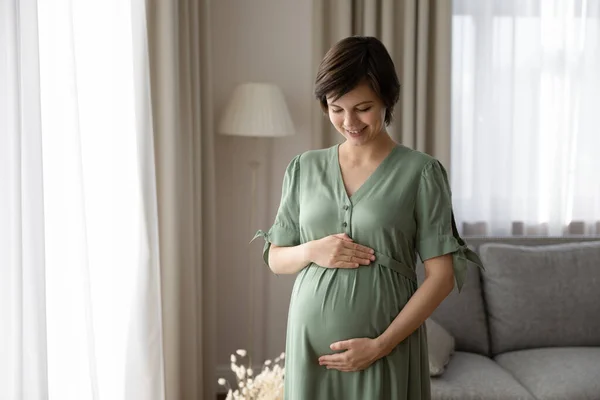 Mujer embarazada sonriente en vestido abrazando el vientre, de pie en casa —  Fotos de Stock