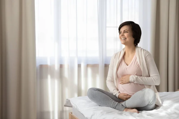 Sorridente donna incinta sognante toccare pancia, seduto sul letto — Foto Stock