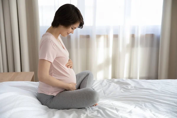 Vista laterale felice donna incinta accarezzando pancia, rilassante in camera da letto — Foto Stock