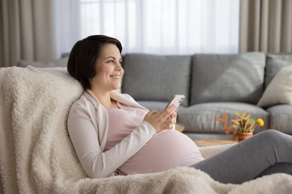 Mulher grávida feliz distraído do smartphone, relaxando na cadeira aconchegante — Fotografia de Stock