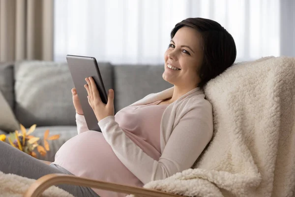 Sorrindo mulher grávida sonhadora usando tablet, se divertindo com o dispositivo — Fotografia de Stock