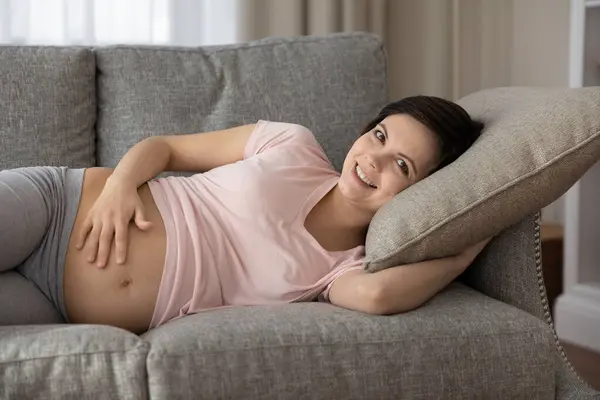 Portrait of happy pregnant woman touching belly, relaxing on couch — Stock Photo, Image