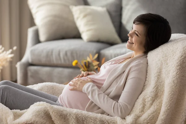 Dreamy smiling pregnant woman touching belly, relaxing in cozy chair — Stock Photo, Image