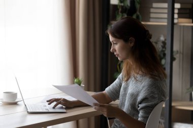 Dizüstü bilgisayarda çalışan, elinde kağıtlar tutan, masada oturan odaklanmış bir kadın.
