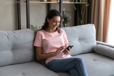 Gülümseyen Arap kadın akıllı telefon kullanıyor, evdeki koltukta oturuyor.