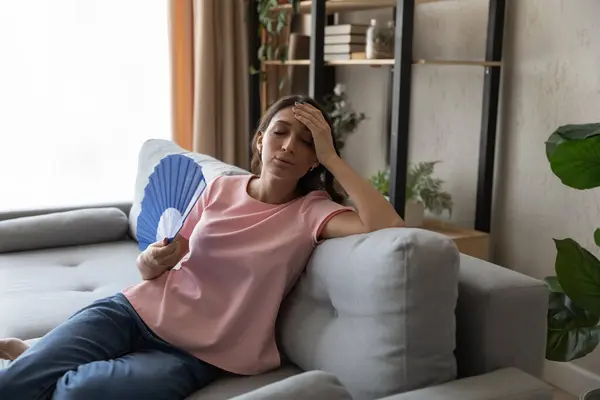 Перегретая арабская женщина машет бумажным вентилятором, сидит на диване — стоковое фото