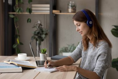 Kulaklıklı gülümseyen kadın not alıyor, internetten ders çalışıyor.