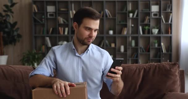 Hombre usando teléfono inteligente mientras se sienta en el sofá con la caja de paquetes — Vídeos de Stock