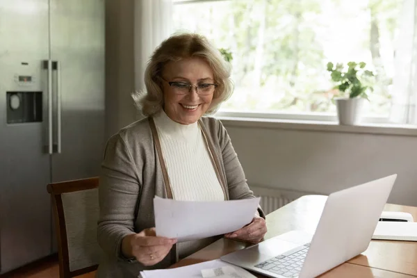 Happy older woman busy working on laptop with paperwork
