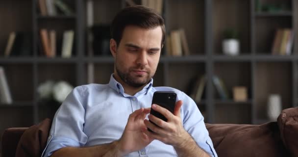 Jovem sorridente satisfeito sente-se no sofá usando o dispositivo de celular — Vídeo de Stock