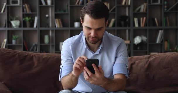 Genç ve yakışıklı bekar adam akıllı telefon cihazıyla koltukta oturuyor. — Stok video