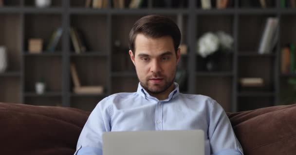 Millennial man zitten op de bank werken vanuit huis met behulp van laptop — Stockvideo