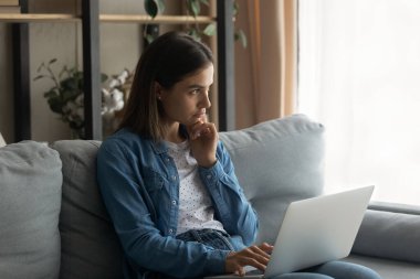 Thoughtful young woman sitting on sofa with computer on laps.